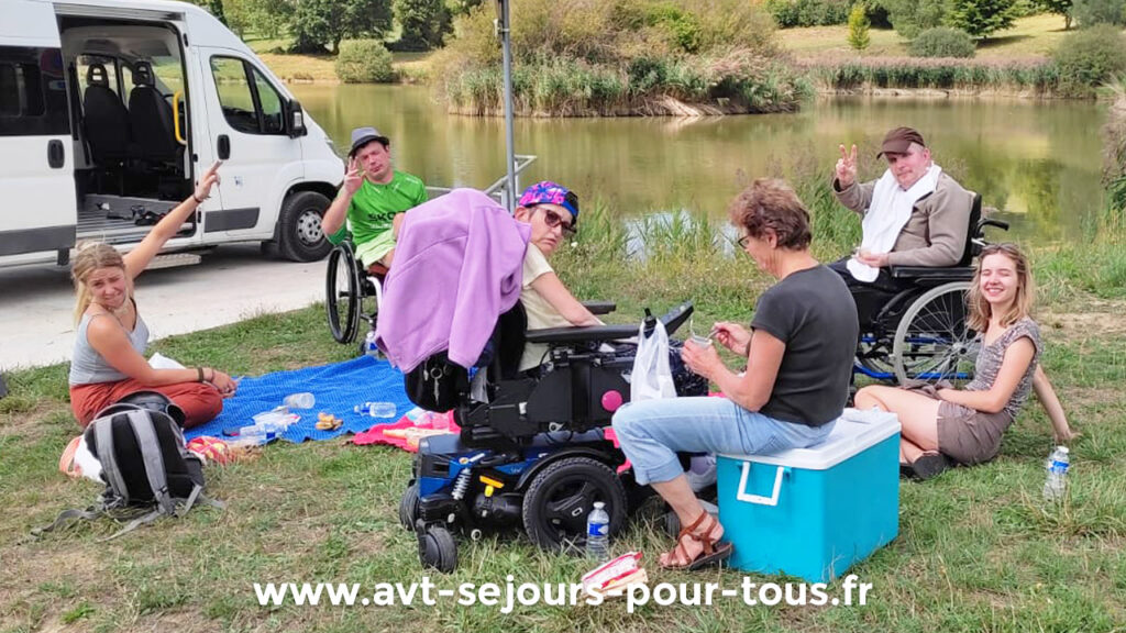 Séjour adapté pmr été à la montagne avec AVT Séjours pour tous. Des vacanciers en fauteuil roulant et leurs accompagnateurs en train de pique-niquer au bord d'un lac.